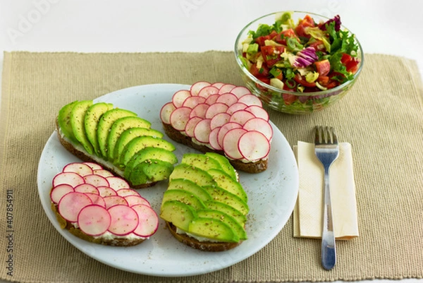 Fototapeta Slices of peasant bread with sliced avocado and radishes on a plate with a salad on the side. 