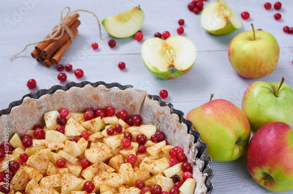 Fototapeta Close up view on apple pie in the
baking dish decorated with fresh apples, cranberry, spices, cinnamon sticks on the gray kitchen background. Autumn tart with raw ingredients