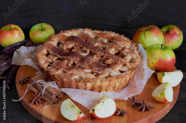 Fototapeta Traditional apple pie with spices on the brown wooden board. Apple pie decorated with sliced fresh apples, carob pods and cinnamon sticks  on the black background. Side view 