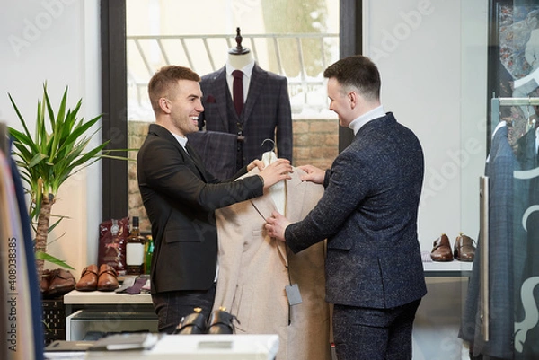 Fototapeta A smiling shop assistant is demonstrating a coat to a man in a white turtleneck sweater and a suit in a clothing store. A male customer and a seller in a boutique.