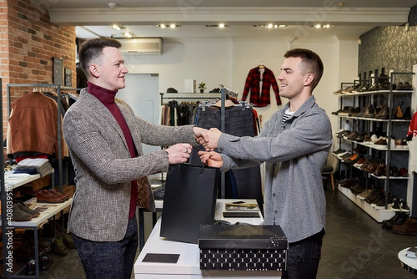 Obraz A happy man is taking his purchase and shaking hands with a smiling shop assistant in a clothing store.