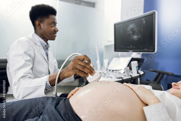 Fototapeta Young professional male African doctor, looking at the screen, while performing ultrasound scan of the abdomen of pregnant Caucasian woman in clinic. Focus on ultrasound transducer
