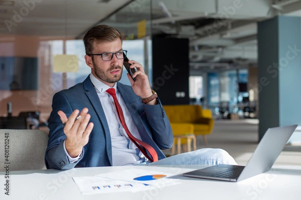 Fototapeta Portrait of young handsome man in eyeglasses talking on the phone with a client while sitting in the office