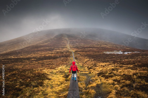 Fototapeta Boy in a red jacket, hiking on wooden path leading through the wicklow mountains, Djouce pek Ireland. Wooden path in foggy mountain landscape, in Autumn. 2019 Irland