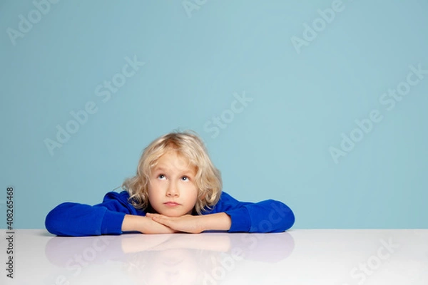 Obraz Dreaming. Happy curly boy isolated on blue studio background. Looks happy, cheerful. Copyspace for ad. Childhood, education, emotions, facial expression concept. Peeking out from behind the table