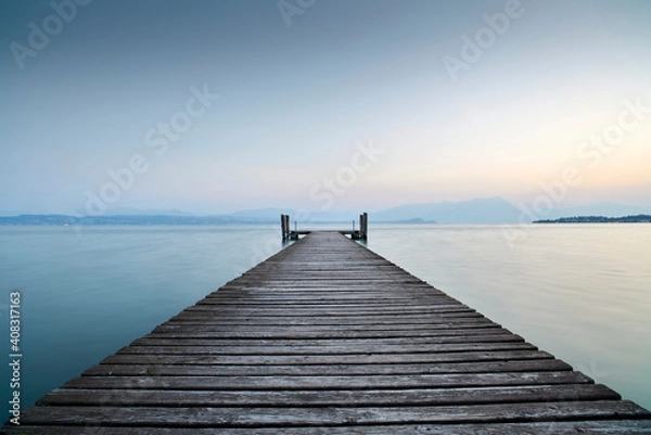 Fototapeta An old wooden pier extends into the clear blue waters of the lake. A solitary path towards the calm and peaceful silence of nature, with the sweet sound of the waves.