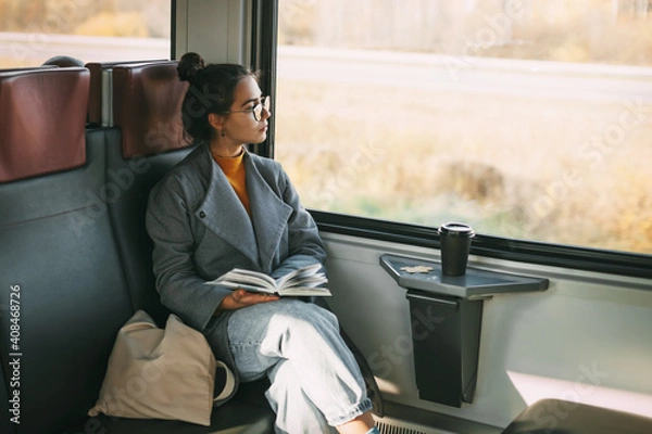 Fototapeta Young beautiful girl on a train reading a book while traveling in a train