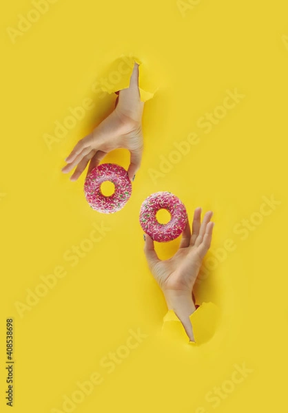 Fototapeta two hands holds delicious pink round glazed donut with sprinkle in torn yellow bright paper hole, vertical popular food blog content, selective focus