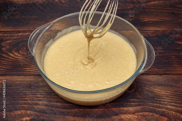 Fototapeta Pasty, liquid dough in a glass bowl with a whisk on a brown wooden table close-up, the concept of making pancakes for breakfast, baking desserts, pie and cake recipe