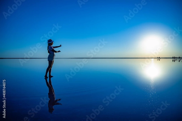 Fototapeta Lovely happy young girl walks and poses in a magical pose along the mirror pink salt lake enjoying the warm evening sun, looking at the fiery sunset and her reflection