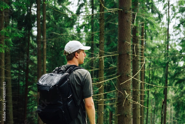 Fototapeta Young male hiker stands with his back in the woods with a backpack on his back and looks away with a serious face. tourist is resting on a hike.