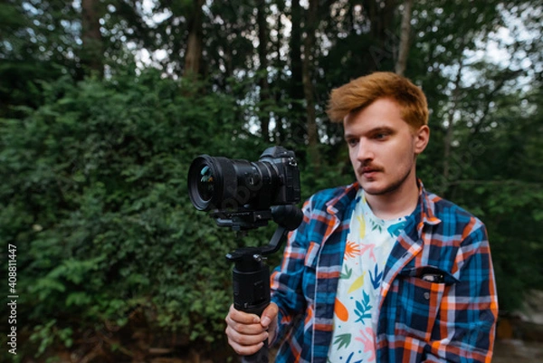 Fototapeta Young man shoots a travel video on camera on a stabilizer with a serious face. Blogger creates video content in nature.