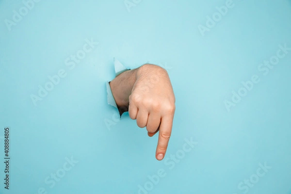 Fototapeta Finger pointing down through torn blue paper wall. Hand gestures of direction.