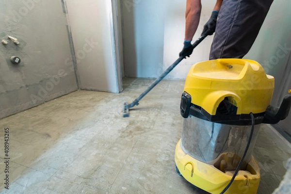 Obraz The builder cleans the floor from dust with a construction vacuum cleaner.