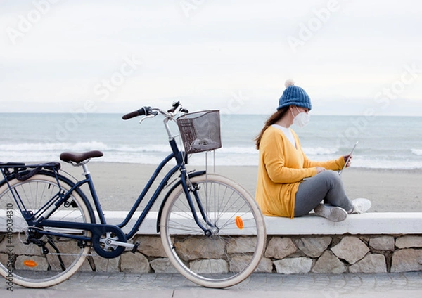Fototapeta A digital marketer is working on a new laptop promotional project while sitting on the seaside promenade with bicycle. The independent woman communicates with clients.