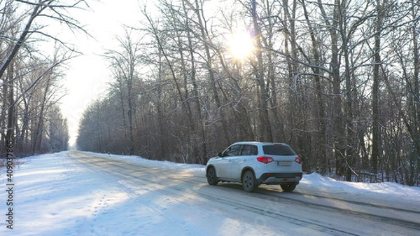 Obraz White car fast riding through snow covered icy road. SUV going at empty countryside route in winter forest on sunny day. Auto moving through scenic landscape way. Travel concept. Top view Drone shot