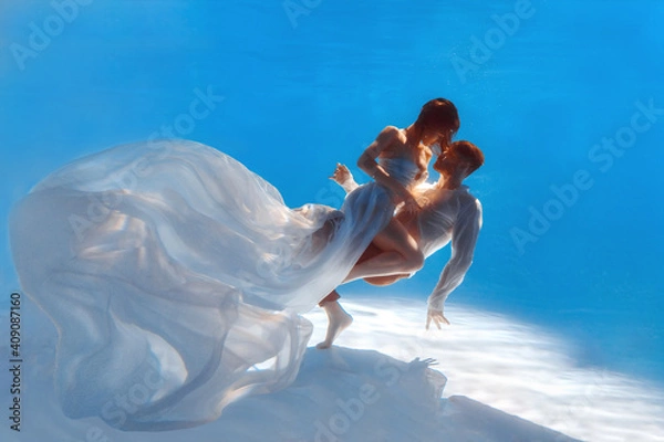 Fototapeta The couple is dancing or hugging in the pool underwater. A girl in a dress with a long train and a guy swim underwater