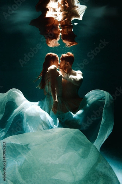Fototapeta The couple is dancing or hugging in the pool underwater. A girl in a dress with a long train and a guy swim underwater