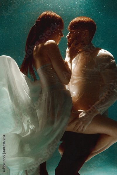 Fototapeta The couple is dancing or hugging in the pool underwater. A girl in a dress with a long train and a guy swim underwater