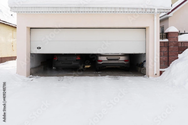 Fototapeta xterior of a garage attached to a house. garage with two cars inside in winter. semi-open sectional doors