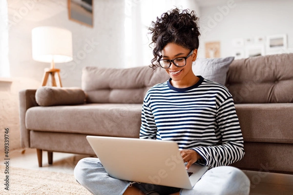 Fototapeta Joyful relaxed ethnic woman using laptop with interest at home