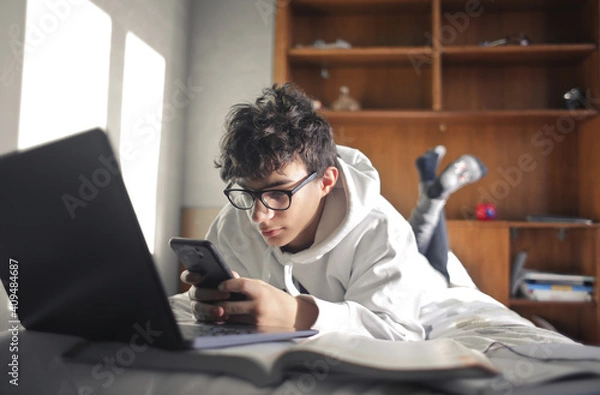 Fototapeta young boy studies lying on the bed using computer and smartphone