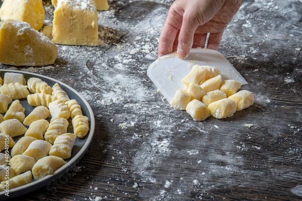 Obraz Gnocchi preparation steps: Woman's hand picking up gluten free handmade mashed potato gnocchi using a scraper. Flour sprinkled on a dark table. Creating authentic fresh pasta dough for Italian dish