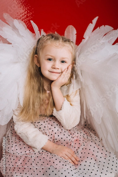 Fototapeta little girl 4 years old in white angel wings on a red latex background in the studio for valentine's day