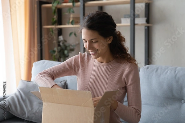 Fototapeta Smiling attractive woman opening big carton box, feeling curious of unpacking internet store order at home. Happy millennial female client satisfied with fast delivery service, online shopping concept