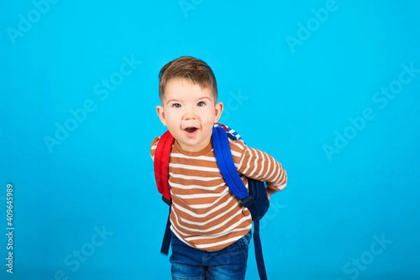 Fototapeta Portrait of surprised happy three year old preschooler with a backpack on blue background. For the first time to school. The concept of developing activities.