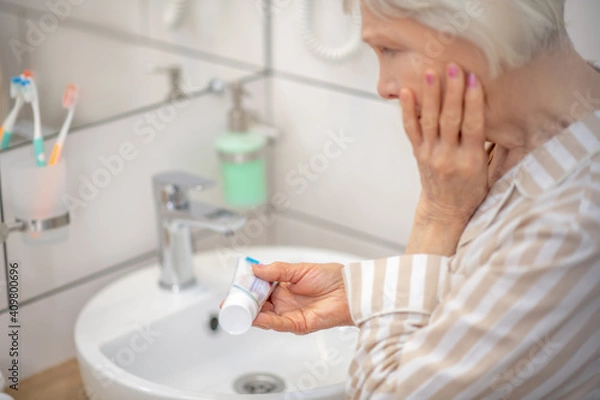 Fototapeta Gray-haired woman in pajama holding a toothpaste and looking at it