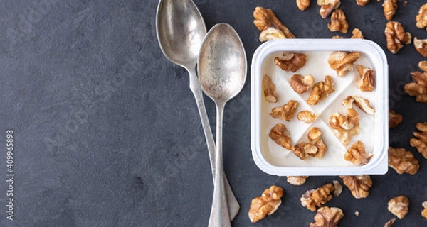 Obraz White yogurt in plastic square cup with marmalade and spoon in with walnuts on gray background. Flat view.