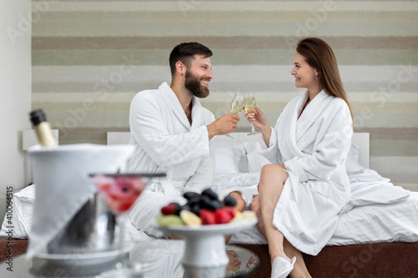 Fototapeta Happy couple a man and a woman in a white coat smile at each other and drink sparkling wine in bed in a hotel room.