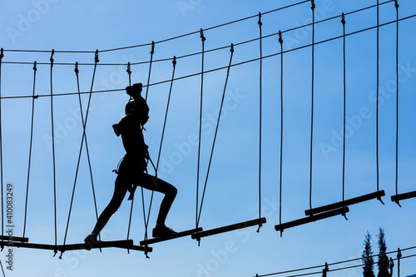 Obraz Silhouette of young girl in helmet climbing on high rope course against blue sky. Wooden bars tied to ropes, part of obstacle course in adventure park