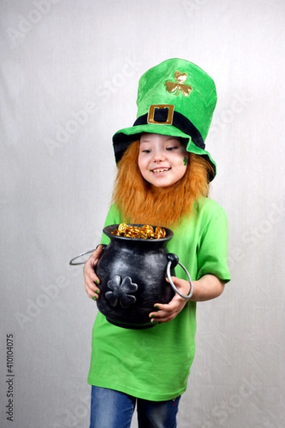 Fototapeta Saint Patricks Day. Funny smiling small girl with decorative red beard, green clover leaf on her cheek and leprechaun hat, dressed in green, looking to cast iron pot, full of leprechaun gold