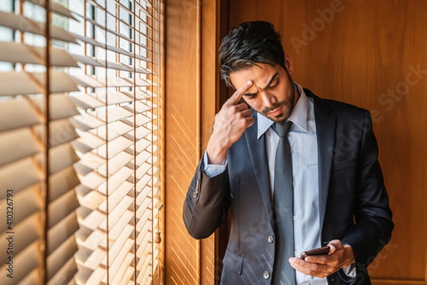 Obraz A businessman standing in front of a window with blinds in a suit feeling upset and frustrated and looking at the mobile phone in his hand.