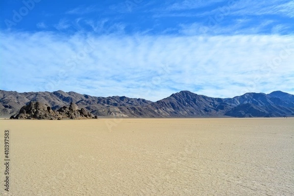 Fototapeta The Grandstand on the Racetrack Playa in the Death Valley National Park, a very dark grey rock surrounded by featureless, light tan-colored clay sediment in a dry lake bed