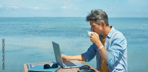 Obraz Pensive man freelancer working on laptop computer at beach by blue sea, drinking coffee. Concept remote work, freelance