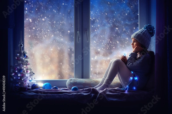 Obraz A girl in a blue hat sits on the windowsill and looks at the snow. Next to her is a Christmas tree, a garland is burning, there is a candle