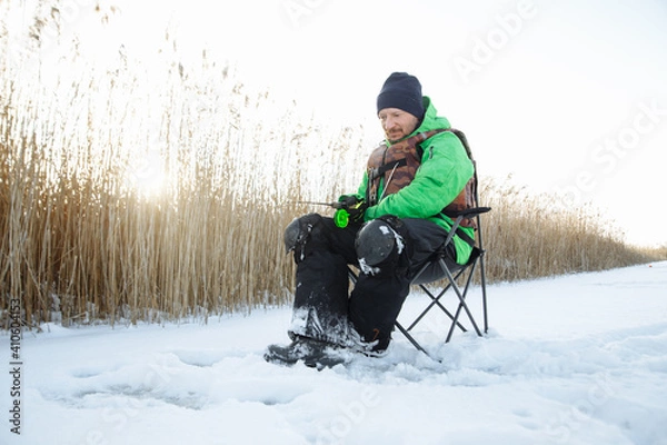 Fototapeta a young man on a winter fisherman sits on a chair and fishes and a fishing rod