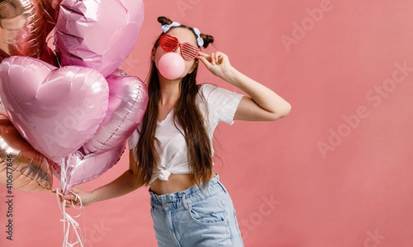 Obraz Young woman dancing, smiling and having fun with bubble gum near big branch of pink heart-shaped ballons. Isolated on pink background.
