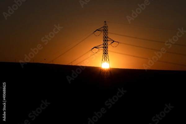 Obraz The sun hiding in the middle of an electricity tower with some mountains in the background against the light. The tower is in the middle of the picture.