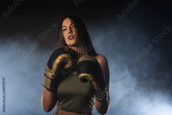 Fototapeta Closeup portrait of a female boxer posing with boxing gloves and looking at the camera with a smokey background