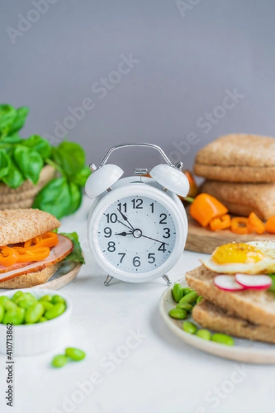 Fototapeta Alarm clock, healthy sandwiches with whole grains bread, microgreens, fresh vegetables Ketogenic diet, intermittent fasting, weight loss. Breakfast and dieting time concept. White table, copy space.