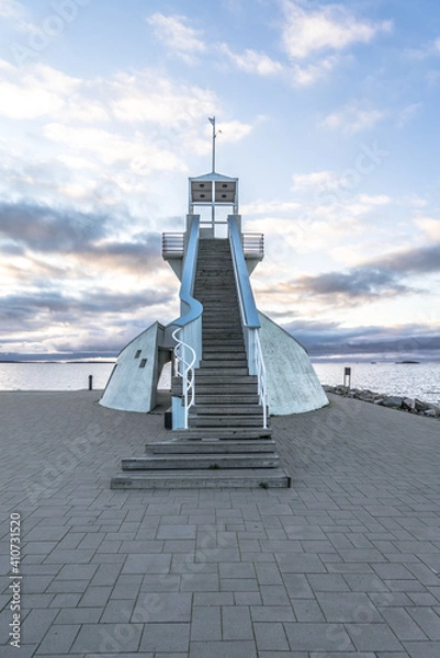 Fototapeta Symmetric staircase at Nallikari lighthouse in Oulu, Finland. Scenic evening skyline over deep blue Baltic sea with stunning clouds at sunset. Maritime tower at the end of stone paved pier-upright