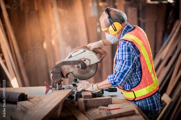 Obraz Asian elderly carpenter craftsman in carpentry shop use circular saws to cut wood board to make furniture. And wear safety equipment at work such as dust masks, gloves, noise-canceling, headphones.