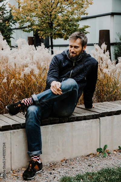 Obraz Handsome middle-aged Caucasian man portraited in the city, sitting on a bench with grey and black hair having a friendly look on his face, wearing a blue jacket and jeans. Fall colourful background.