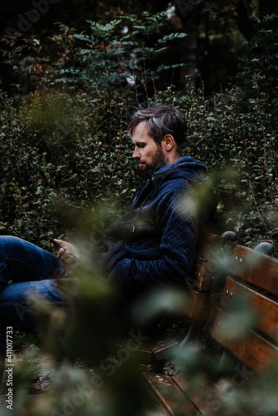Obraz Handsome middle-aged Caucasian bearded man portrayed in the city, sitting on a park bench, holding a cell phone, with grey and black hair having a friendly look on his face, wearing city clothes.