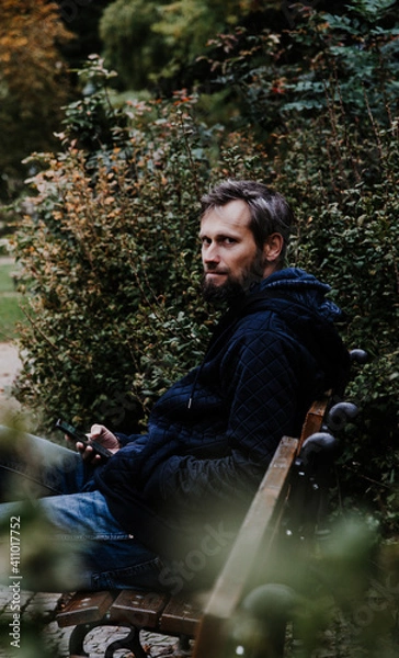 Obraz Handsome middle-aged Caucasian bearded man portrayed in the city, sitting on a park bench, holding a cell phone, with grey and black hair having a friendly look on his face, wearing city clothes.