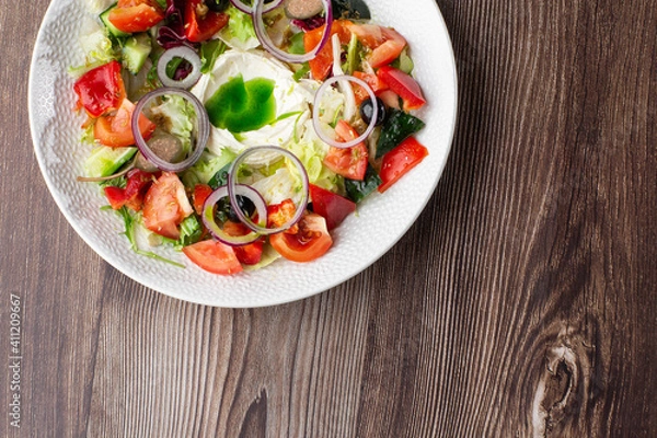 Fototapeta Greek salad with fresh vegetables: tomato, cucumber, red bel pepper, lettuce, onion, olives and cheese. Top view close-up on a white round plate on a wooden background with a copy space. Healthy food.